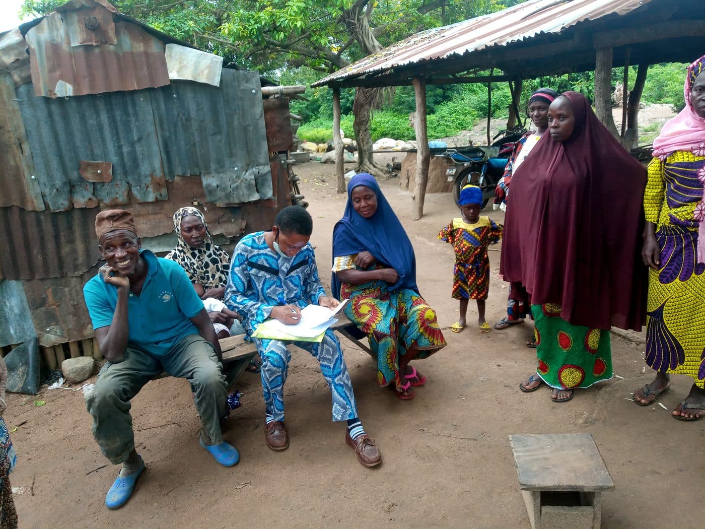 A member of our field team recording answers on paper, seated on a bench with community members in a household compound